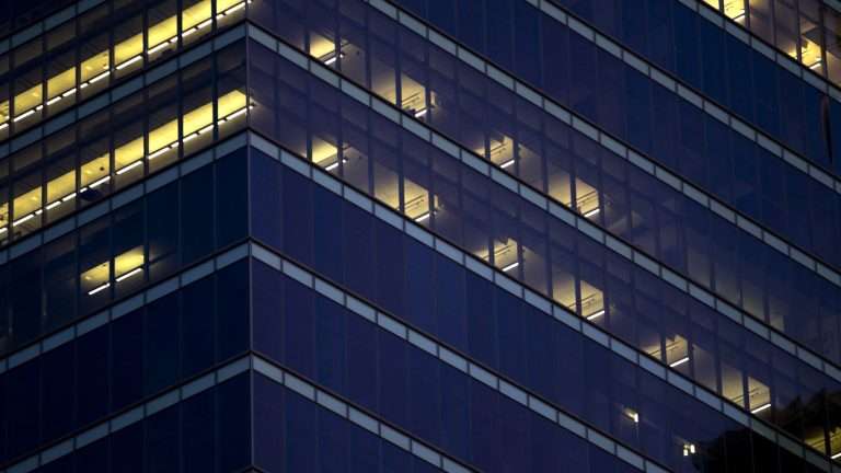 The corner of a dark office building at night with a few floors lit